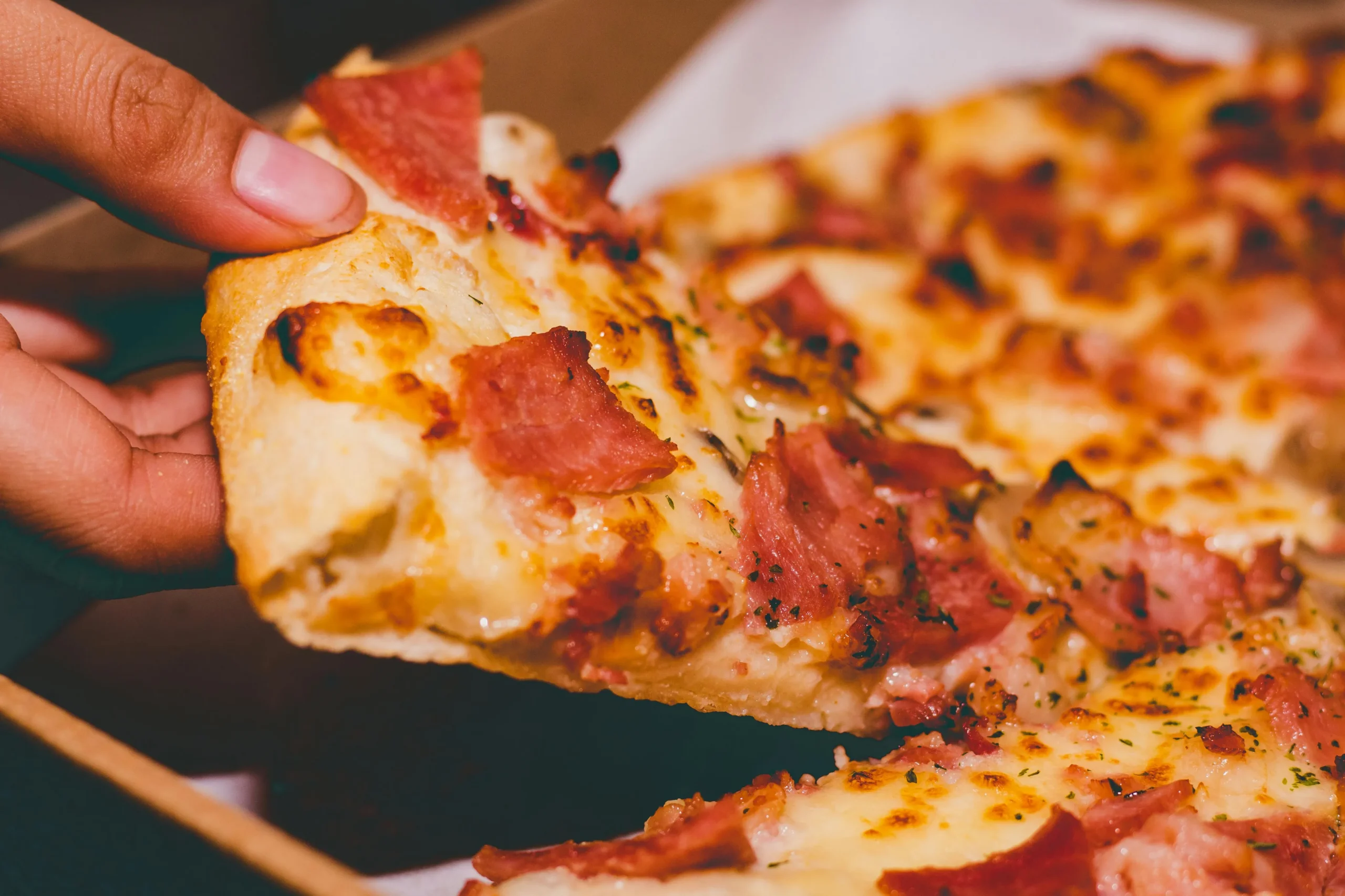 Hand lifting a slice of cheesy pizza topped with ham and herbs from a box scaled