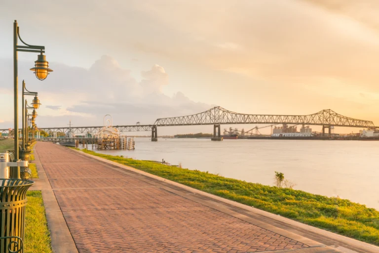 Paved riverfront promenade at sunset featuring street lamps overlooking a cantilever bridge over the water