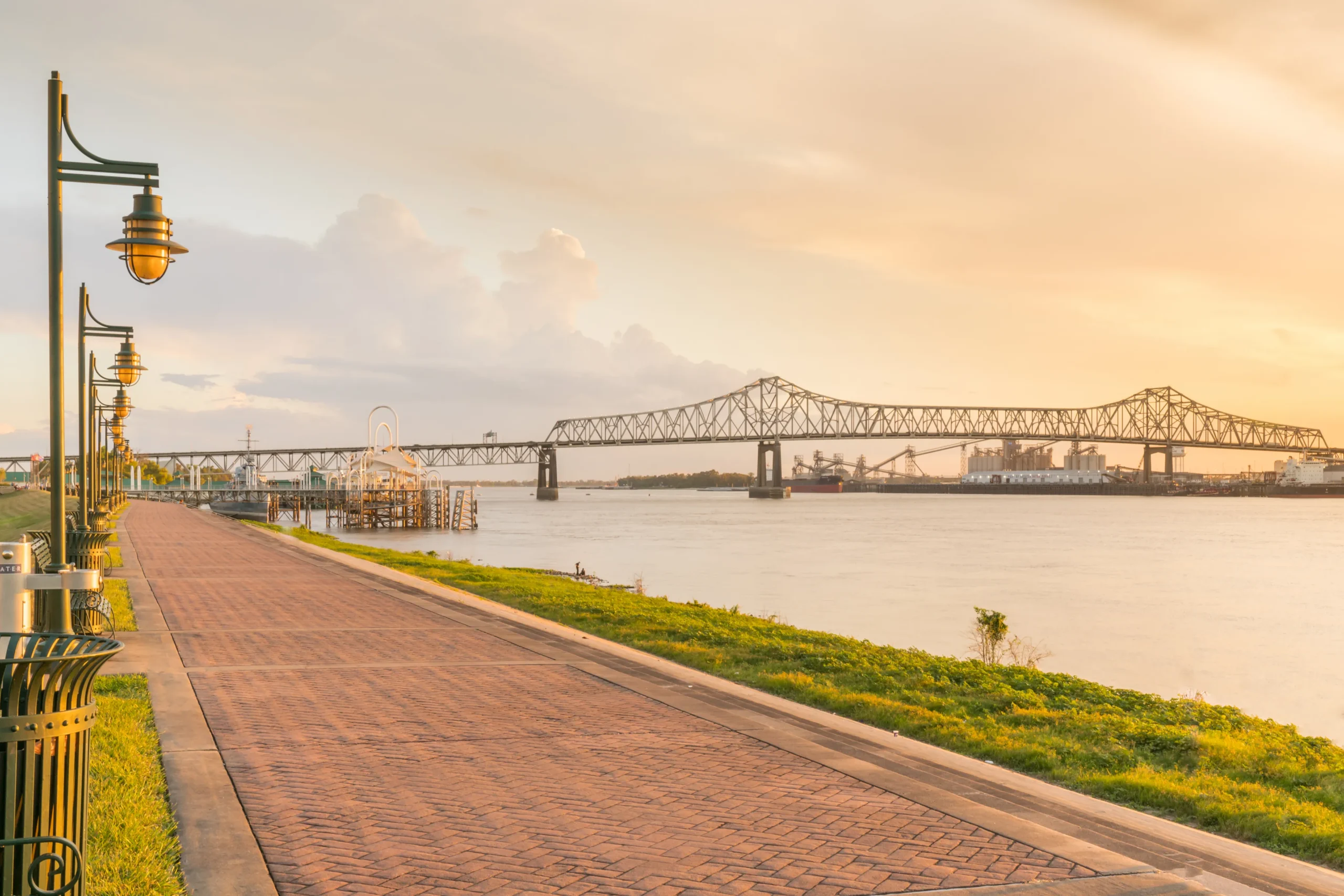 Paved riverfront promenade at sunset featuring street lamps overlooking a cantilever bridge over the water scaled