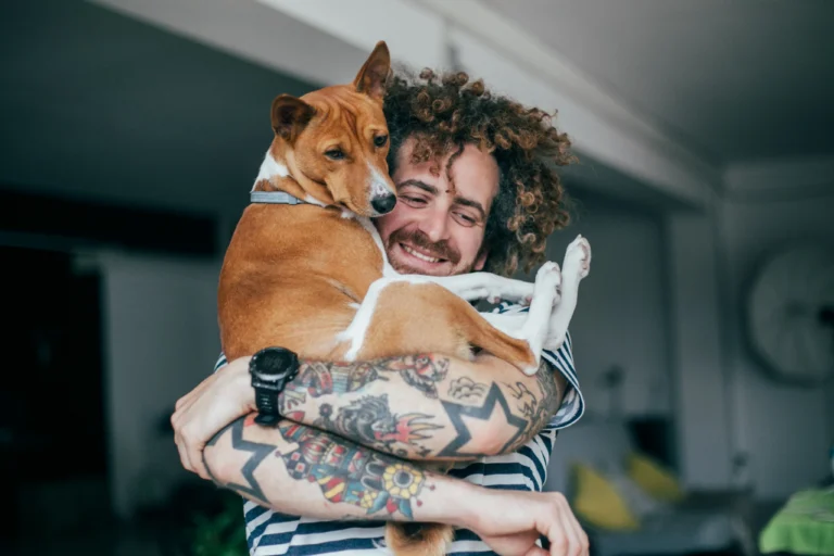 Tattooed man with curly hair smiling while holding a brown and white Basenji dog indoors