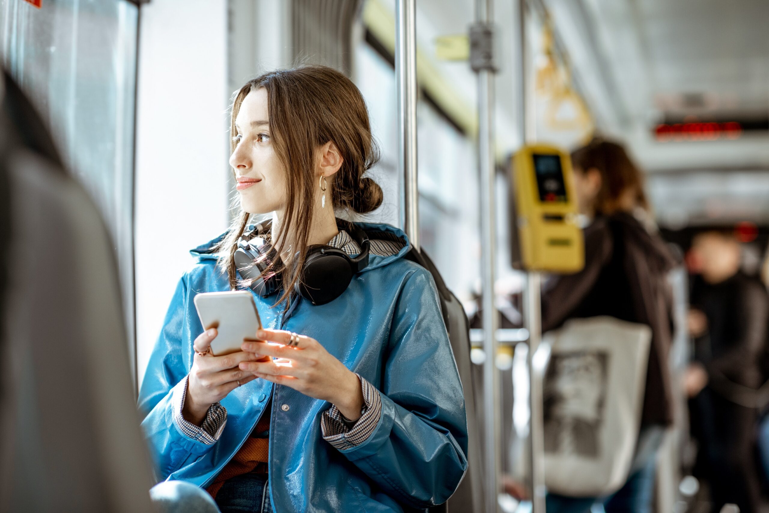 Young woman in a blue jacket looking out the window while holding a smartphone on public transit or a tram scaled